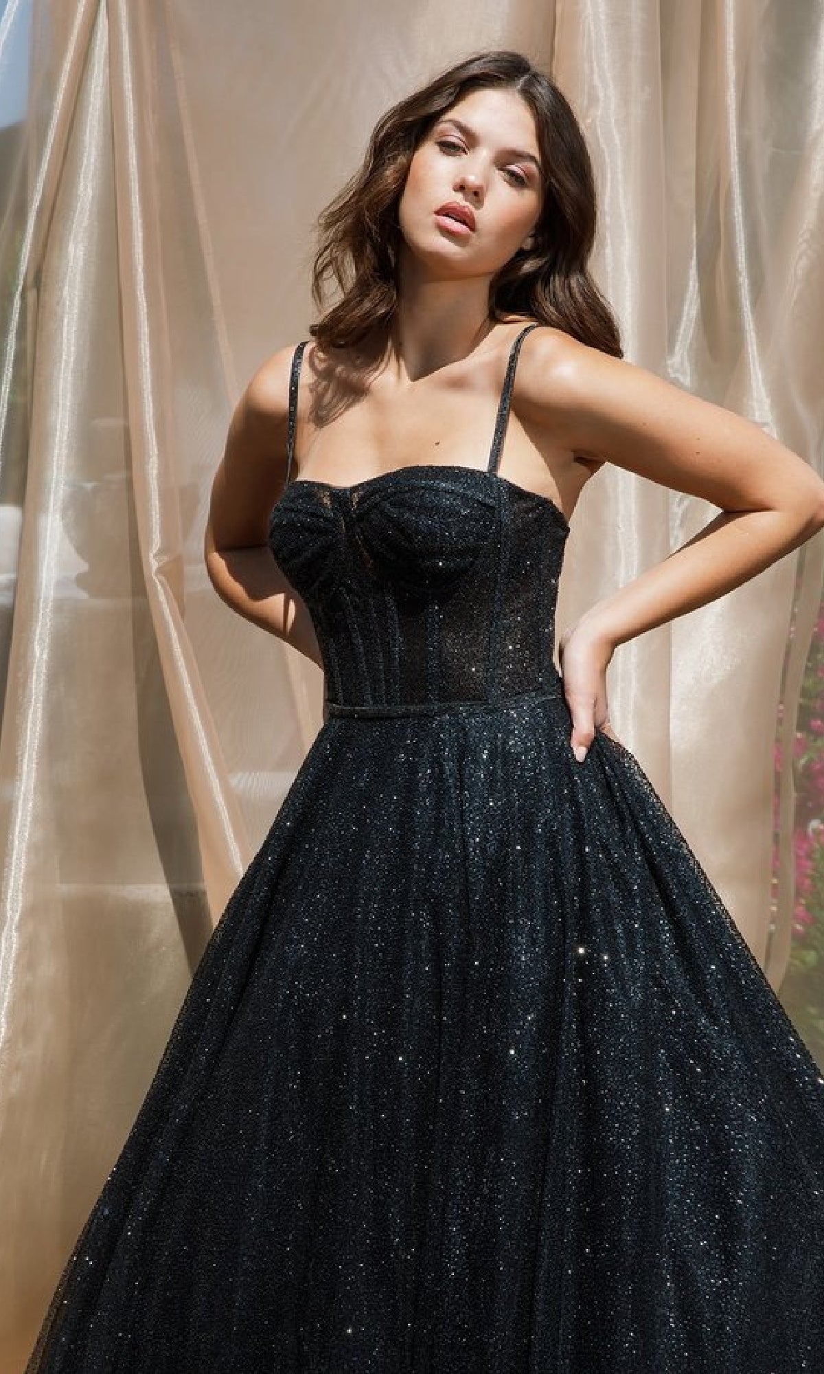A woman stands before sheer curtains wearing the Sheer-Corset Glitter Prom Ball Gown: Eureka 9900—a sleeveless, sparkling black A-line dress with thin straps and a fitted bodice.